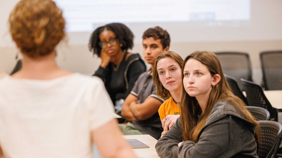 Students attentively listen to a teacher in a classroom setting.